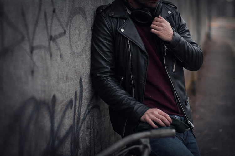 Person wearing a black leather jacket leaning against a graffiti-covered wall.